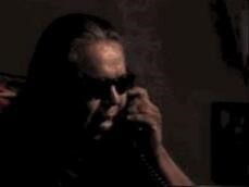 A person wearing sunglasses speaks on a corded phone in a dimly lit room.