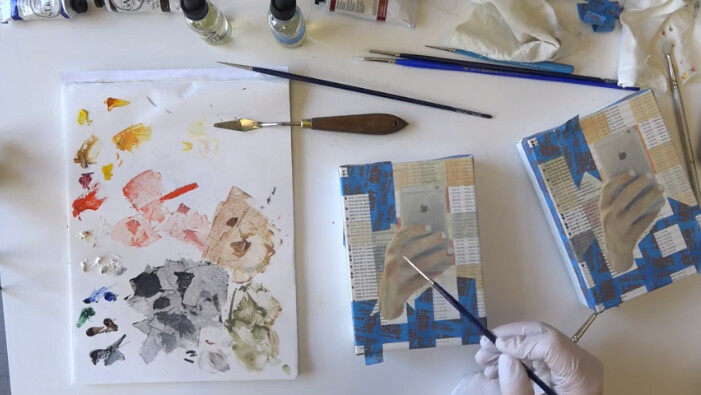 Artist wearing gloves paints a small canvas showing a taped reference image of a hand holding a phone.