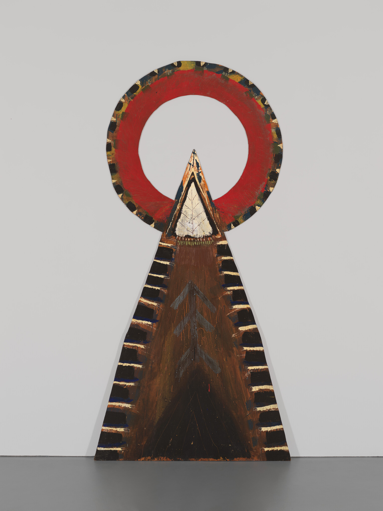 A tall triangular wooden sculpture painted with dark stripes and topped by a large red circular halo.