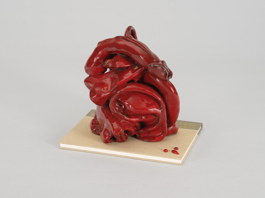 A glossy red abstract ceramic sculpture sits on a closed book with three small red beads.