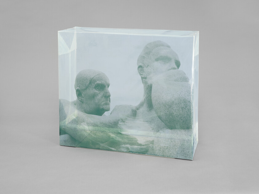 Two large weathered stone heads are encased in a translucent rectangular block on a plain surface.