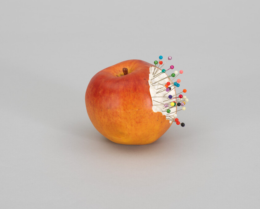 A bitten apple reveals a white pincushion with many colorful sewing pins stuck into it.