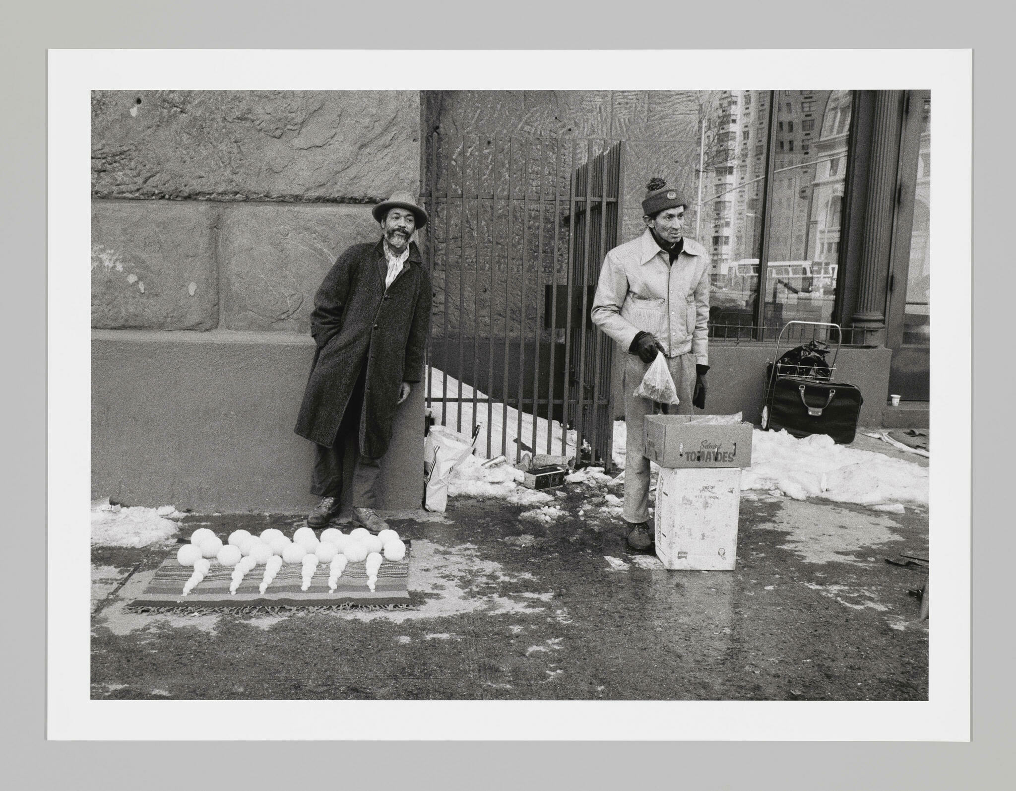 David Hammons, a bearded Black man in a long coat and fedora, stands behind a blanket displaying snowballs organized by size. A person with light skin in a beanie and jacket stands nearby behind stacked boxes.