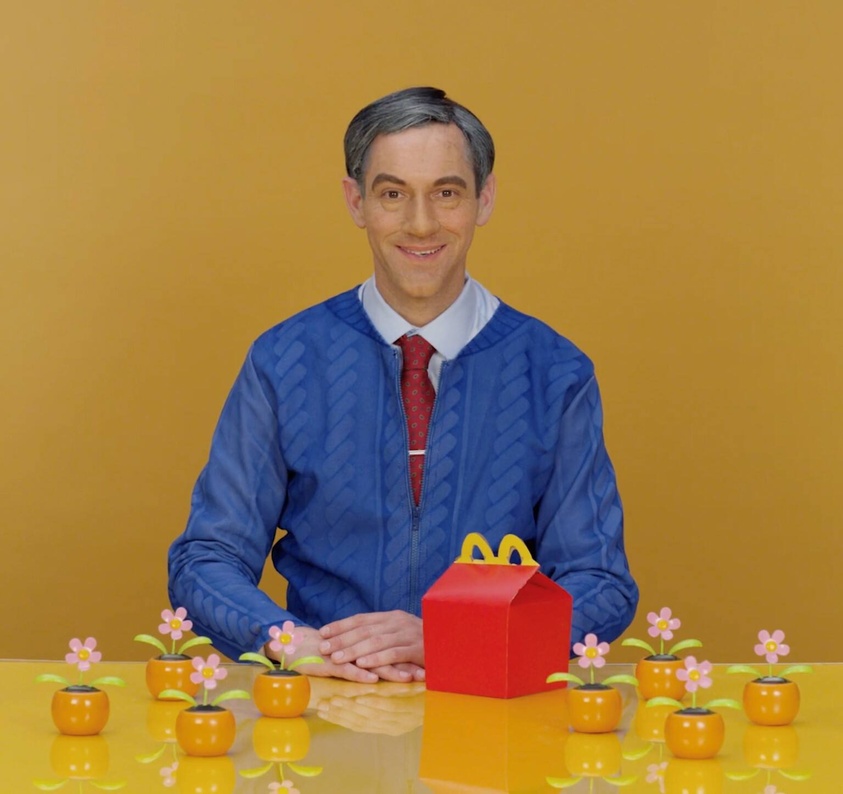 Still of a Mr. Rogers lookalike in a blue cardigan in front of an orange wall at an orange table with flowers and a McDonald’s Happy Meal