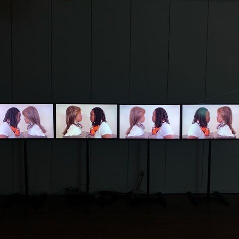 A series of monitors arranged side by side displaying a continuous loop of two women facing each other in conversation, with one woman's back to the viewer on the left and the other woman facing her, visible in profile. The background is a plain, dark wall, and the cables of the monitors are visible on the floor.