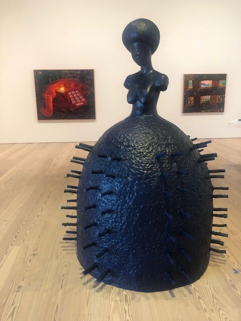 Large dark sculpture of a female bust atop a rounded, nail-studded body displayed in a gallery.