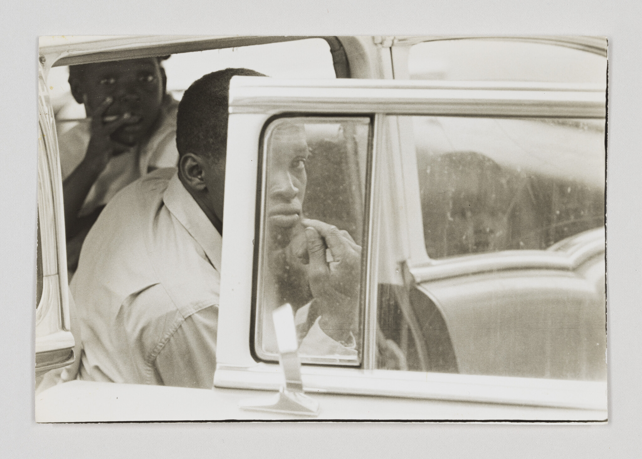 Two men sit in a car backseat, one staring thoughtfully out the window with his hand near his mouth.
