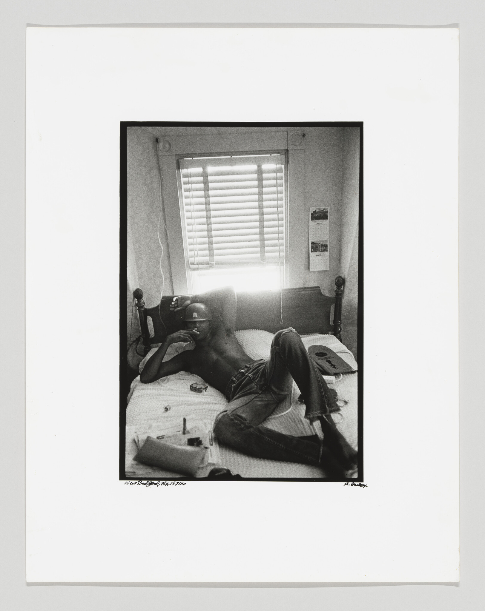 A shirtless man lies on a bed smoking a cigarette beneath a bright window with blinds.