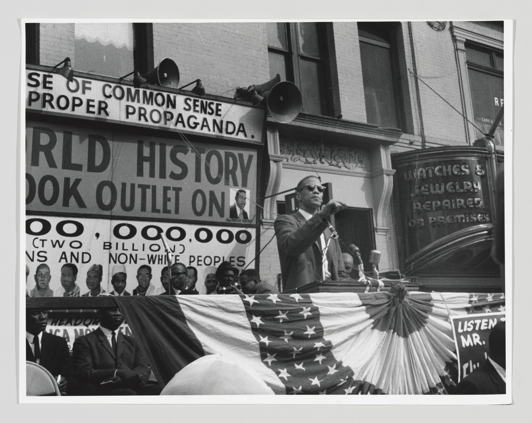 A man wearing sunglasses speaks from a decorated podium to a crowd during an outdoor rally.