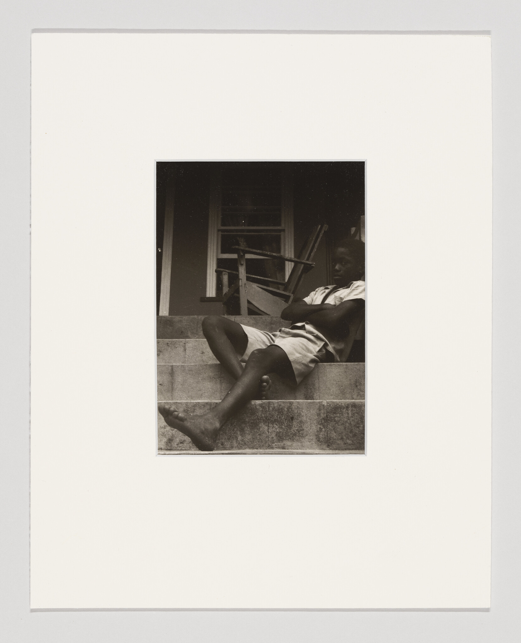 A barefoot boy sits with crossed arms on concrete front steps beside a porch chair.