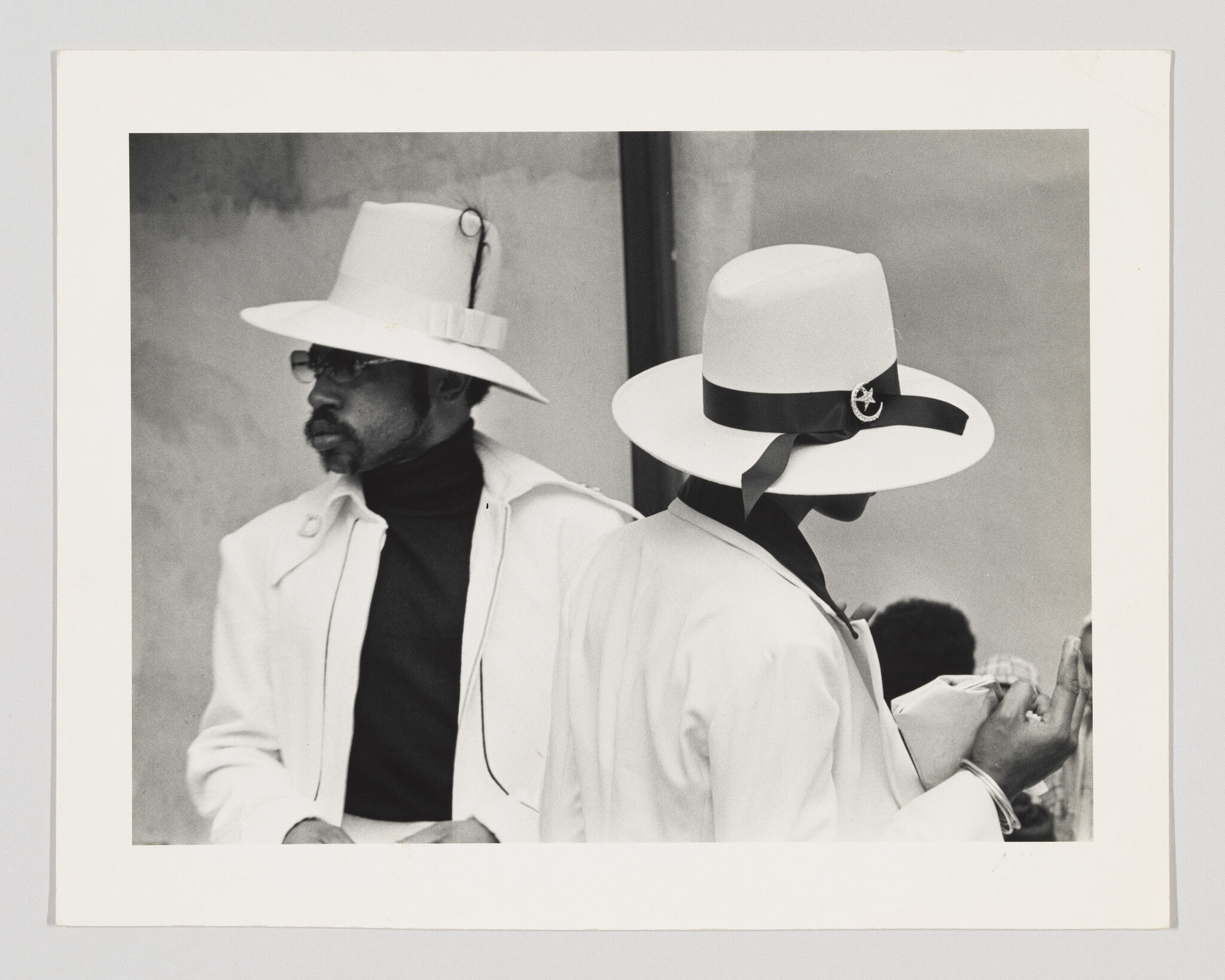 Two people in white hats and jackets stand back-to-back, one holding a clutch and looking at their hand.