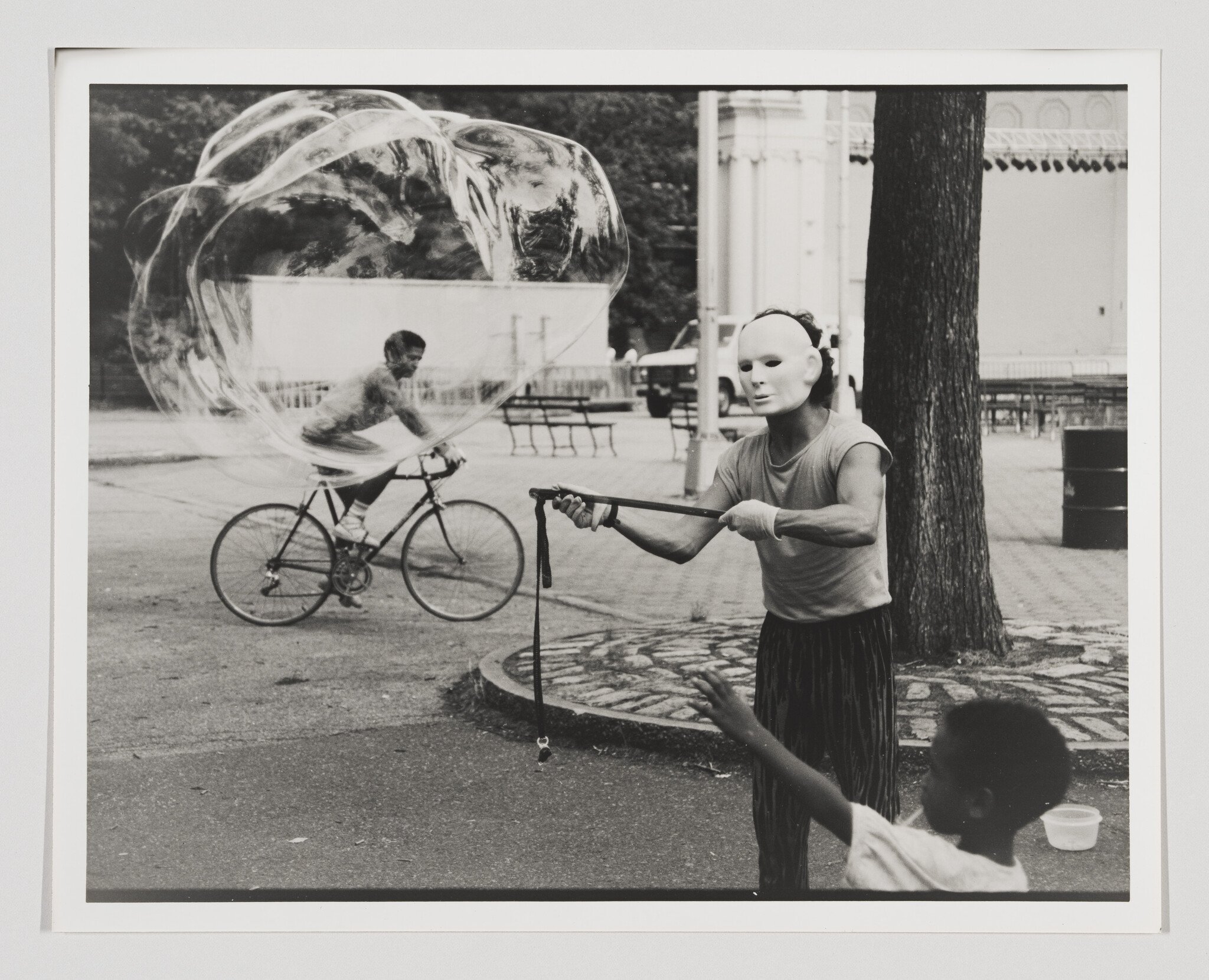 A masked performer creates a giant soap bubble while a child reaches and a bicyclist rides through it.