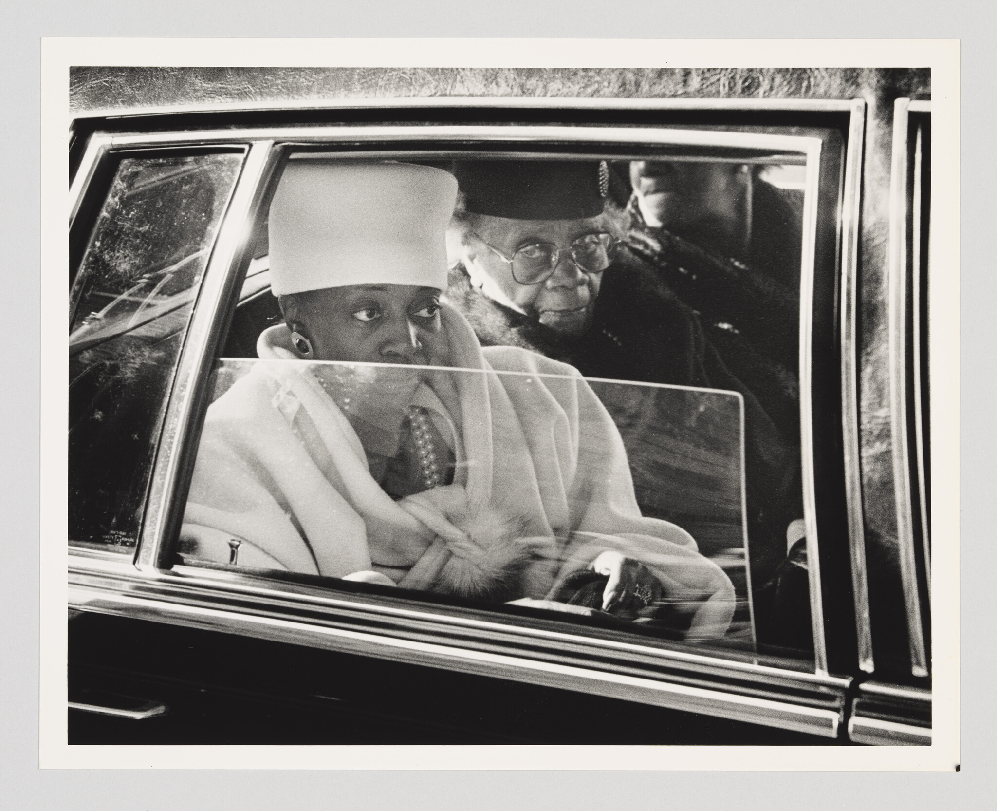 A woman in a tall white hat and pearls sits in a car looking pensively out the window.