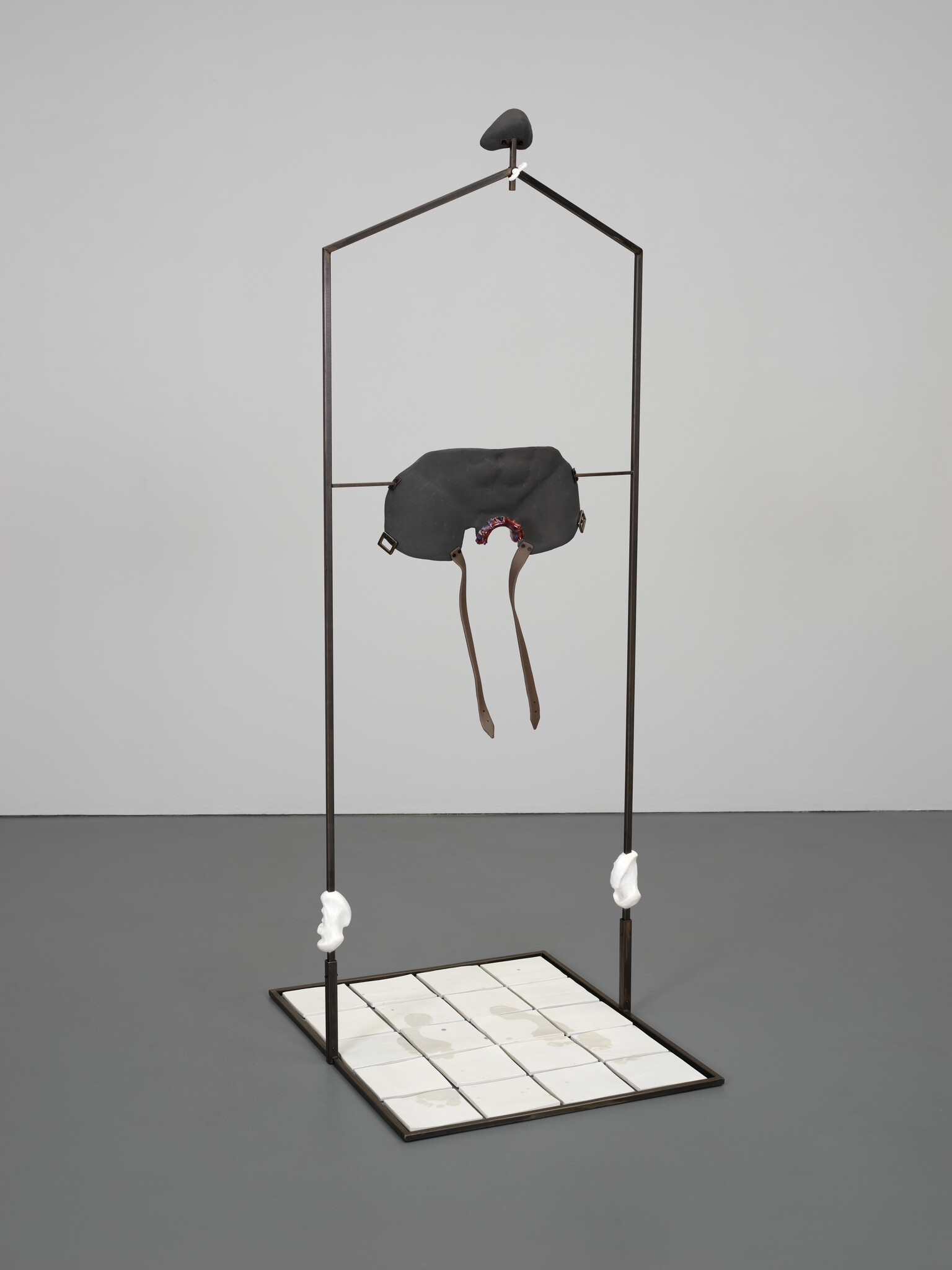 A tall metal frame supports a suspended black mask with dangling leather straps above a tiled base.