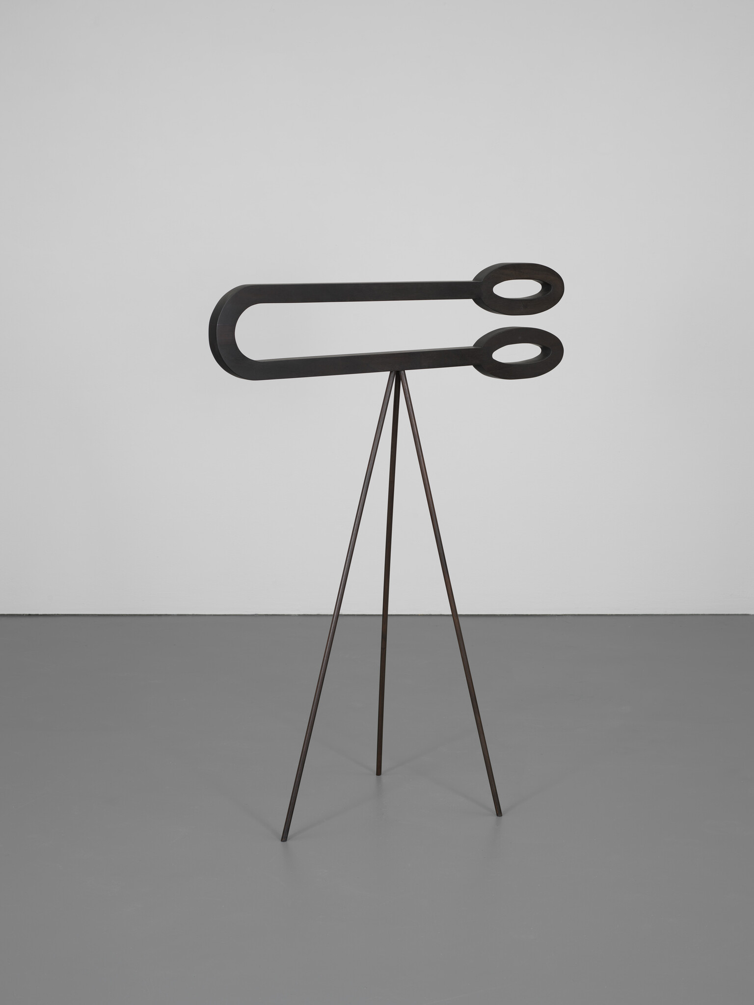 A minimalist black metal sculpture with two elongated oval loops balanced on a three-legged stand.
