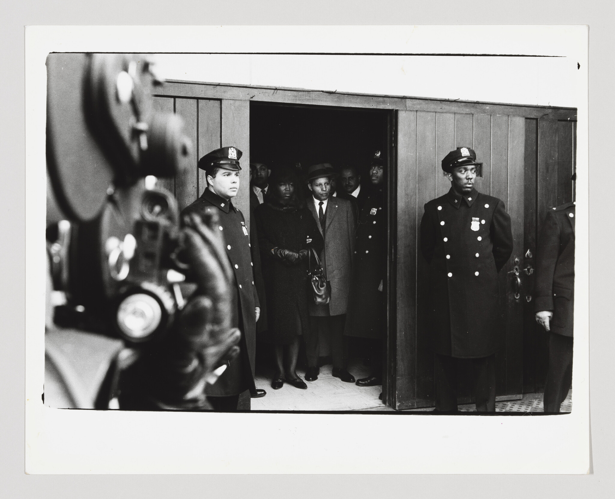 Police officers stand guard at a doorway while a man and woman exit as a film camera films.