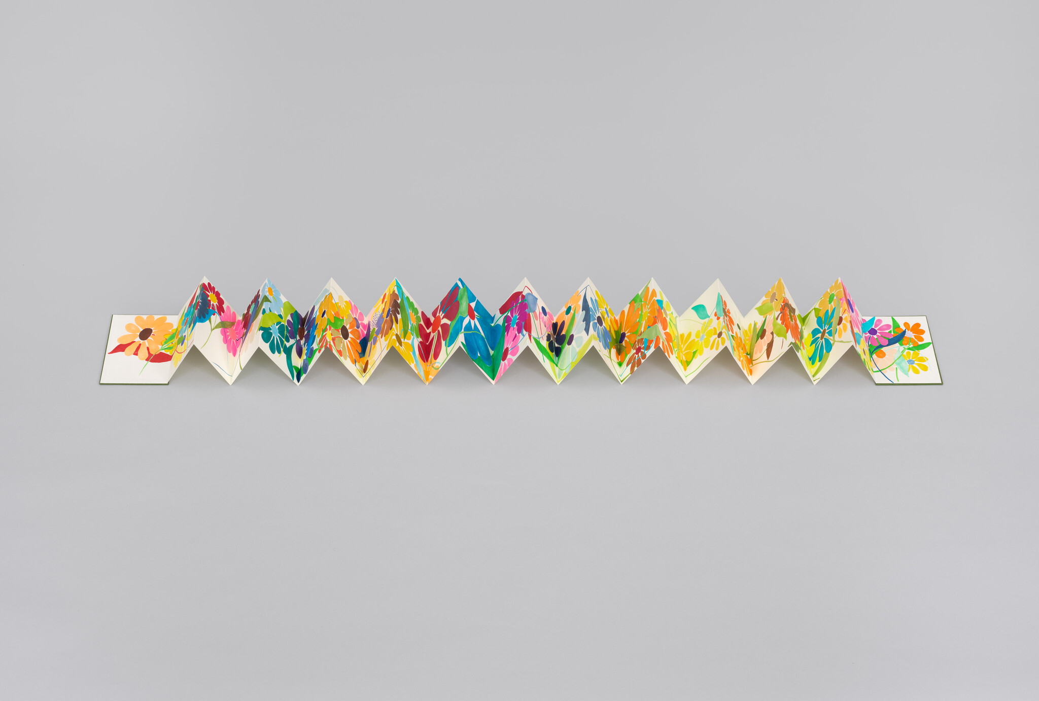 A long accordion-folded paper displays colorful hand-painted flowers in a zigzag across a gray background.