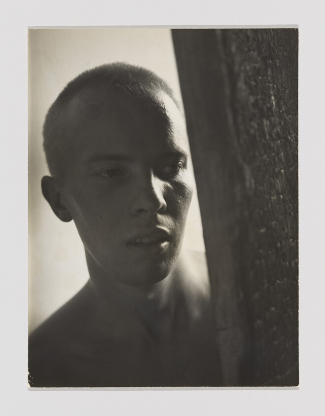Young man with shaved head peers around a wooden post, face partly in shadow.