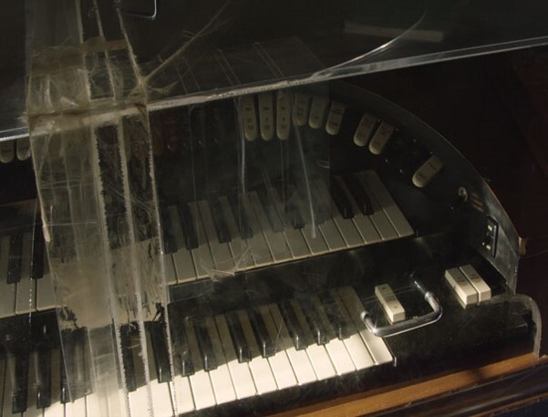 Worn organ keyboard covered in dusty clear plastic with pull-stop knobs above keys.