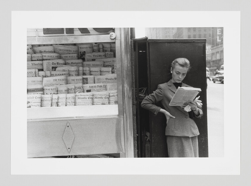 A woman stands by a newsstand on a city sidewalk reading a folded newspaper.