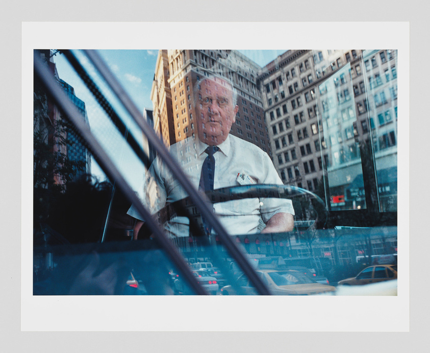 A photograph of a city scene reflected on a car window, superimposing the image of a man in a white shirt and tie over the urban backdrop. The man appears to be sitting inside the vehicle, looking directly at the camera, while the reflection shows buildings, blue sky, and other cars on the street.