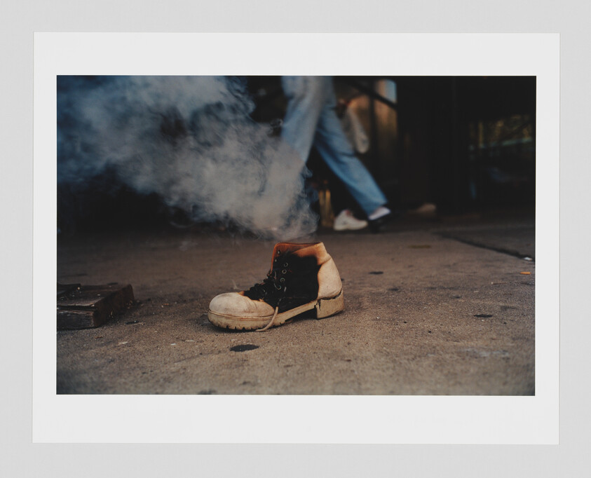 A single worn work boot sits on pavement with smoke rising and a person walking past.