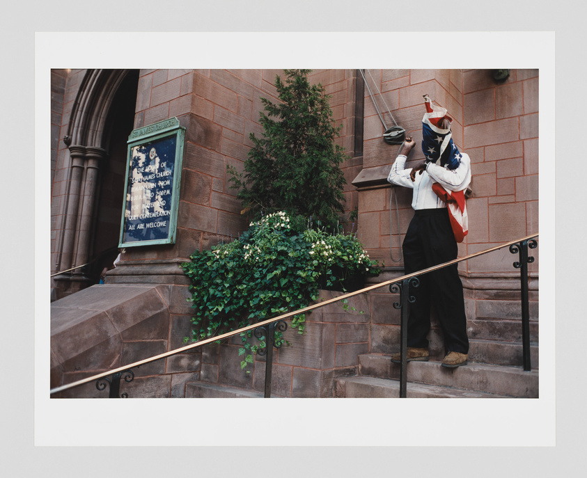 A person dressed in a white blouse and black trousers stands on the steps of a church, adjusting a large American flag over their face. To the left, a sign reads "St. Ann's Church" with additional text below. The scene is framed by lush greenery and the brownstone architecture of the church.