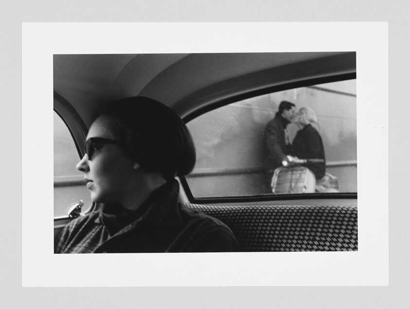 Woman wearing sunglasses sits in a car looking out the window while a couple kisses outside.