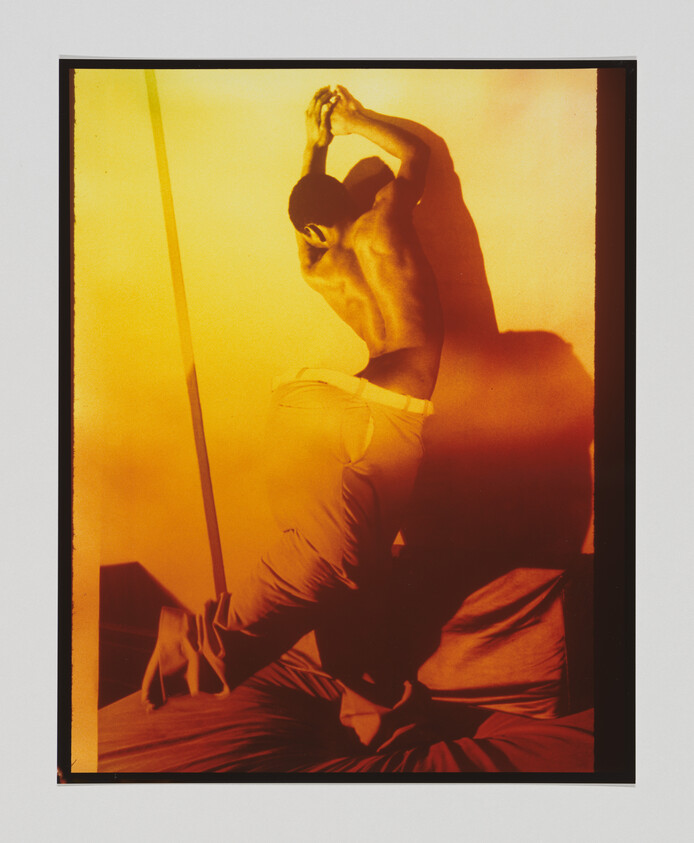 Shirtless man jumps on a bed with arms raised, casting a large shadow on the wall.