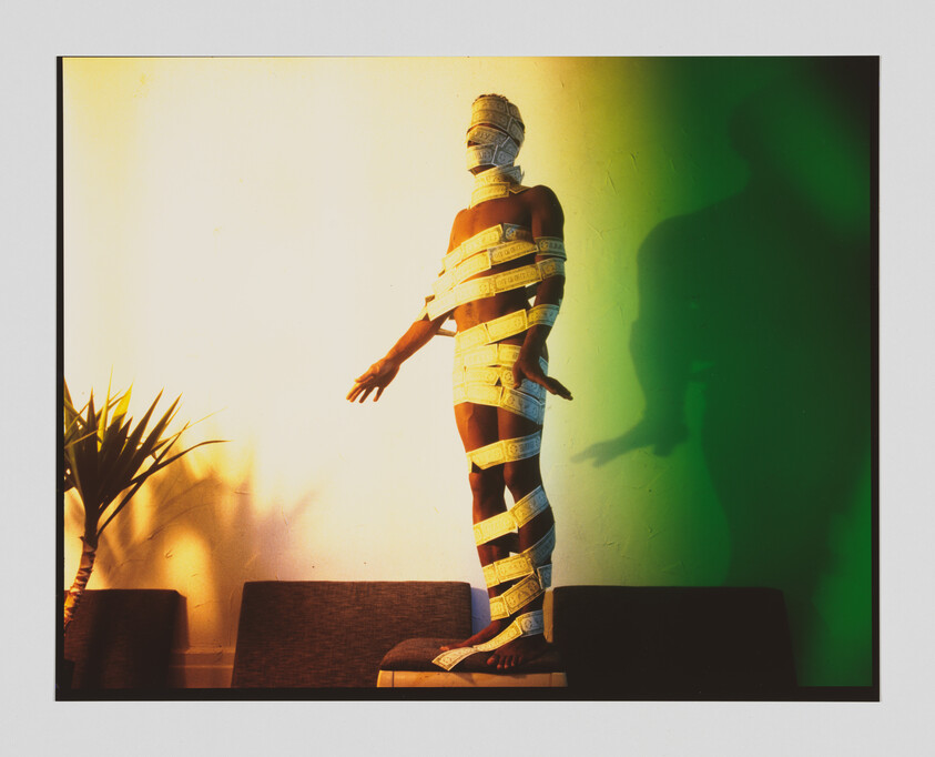 A person stands on a couch wrapped head-to-toe in measuring tape, arms outstretched.