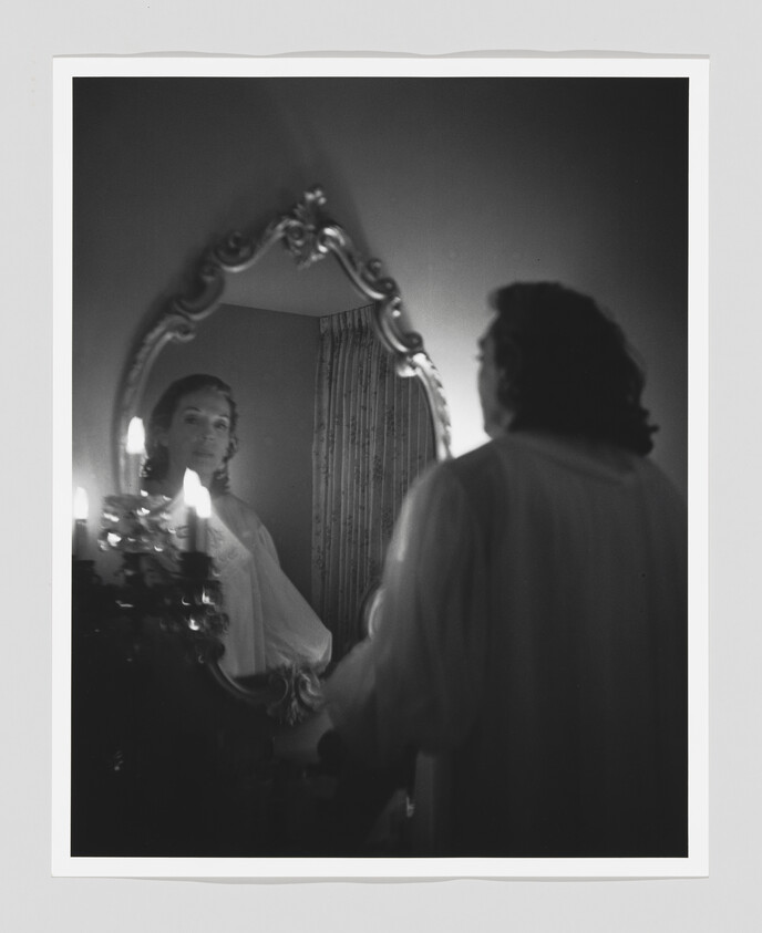 A woman in a nightgown gazes at her reflection in an ornate mirror by candlelight.