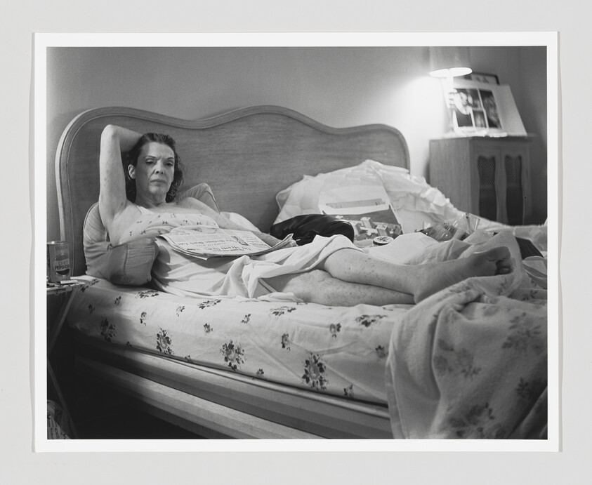 Woman reclining on a bed with a newspaper across her lap, looking toward the camera.