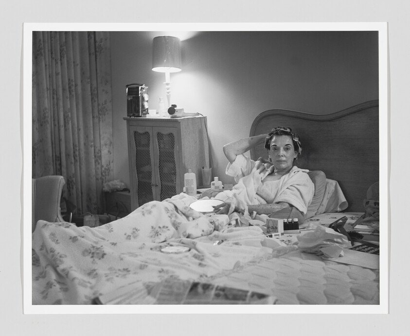 Woman sitting up in bed wearing hair curlers and surrounded by medication and bedside items.