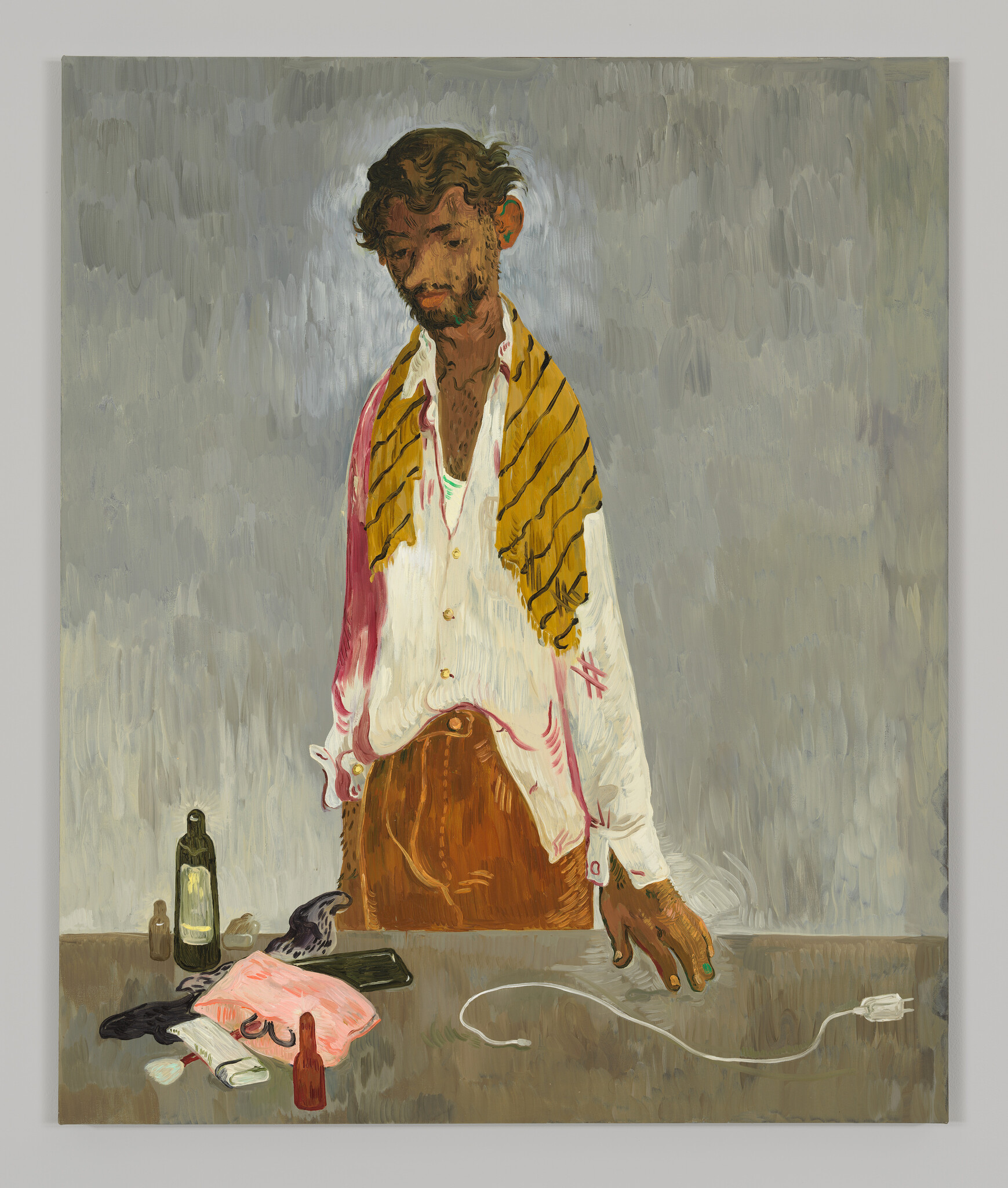 A short haired man with brown skin and a scruffy beard stands over a charger and other small belongings. His shirt is slouchy with one sleeve stained red.