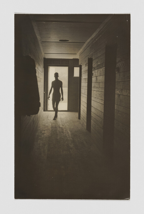 A silhouetted person walks down a dim wooden hallway toward a bright doorway.