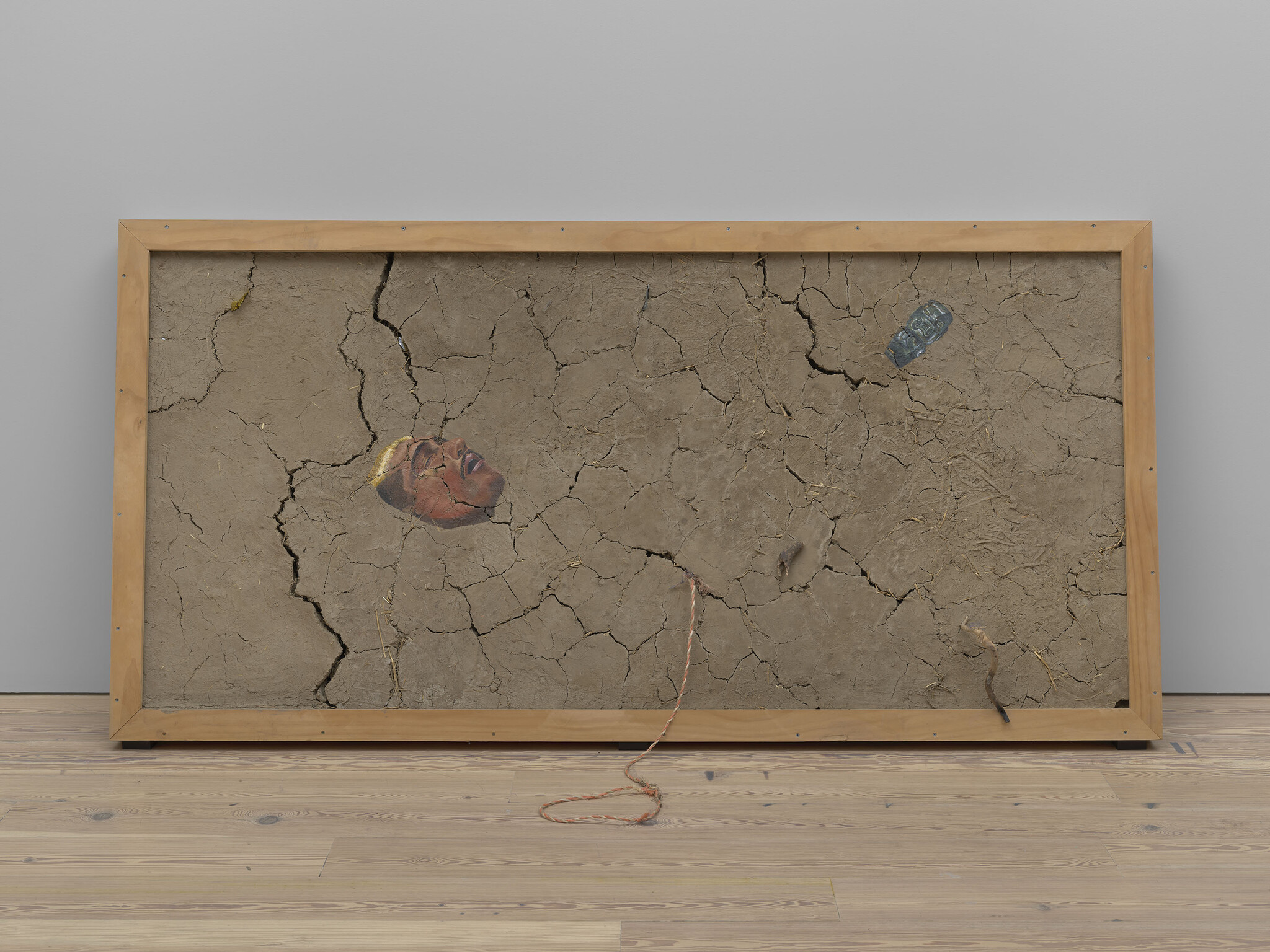 Large wooden frame holds cracked dry mud with a painted human face, rope, and plastic bottle.