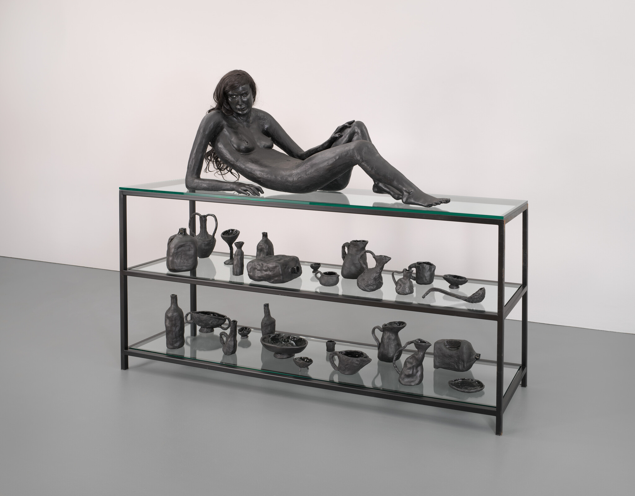 Reclining dark bronze female sculpture rests atop a glass display shelf filled with small vessels.