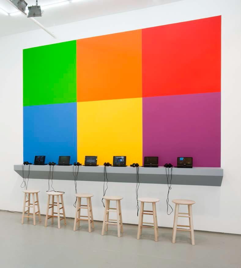 A colorful art installation with a large, pixelated mural of six squares in bright green, orange, blue, red, yellow, and purple. Below the mural, a gray counter holds several laptops with headphones, and wooden stools are placed in front of the counter.