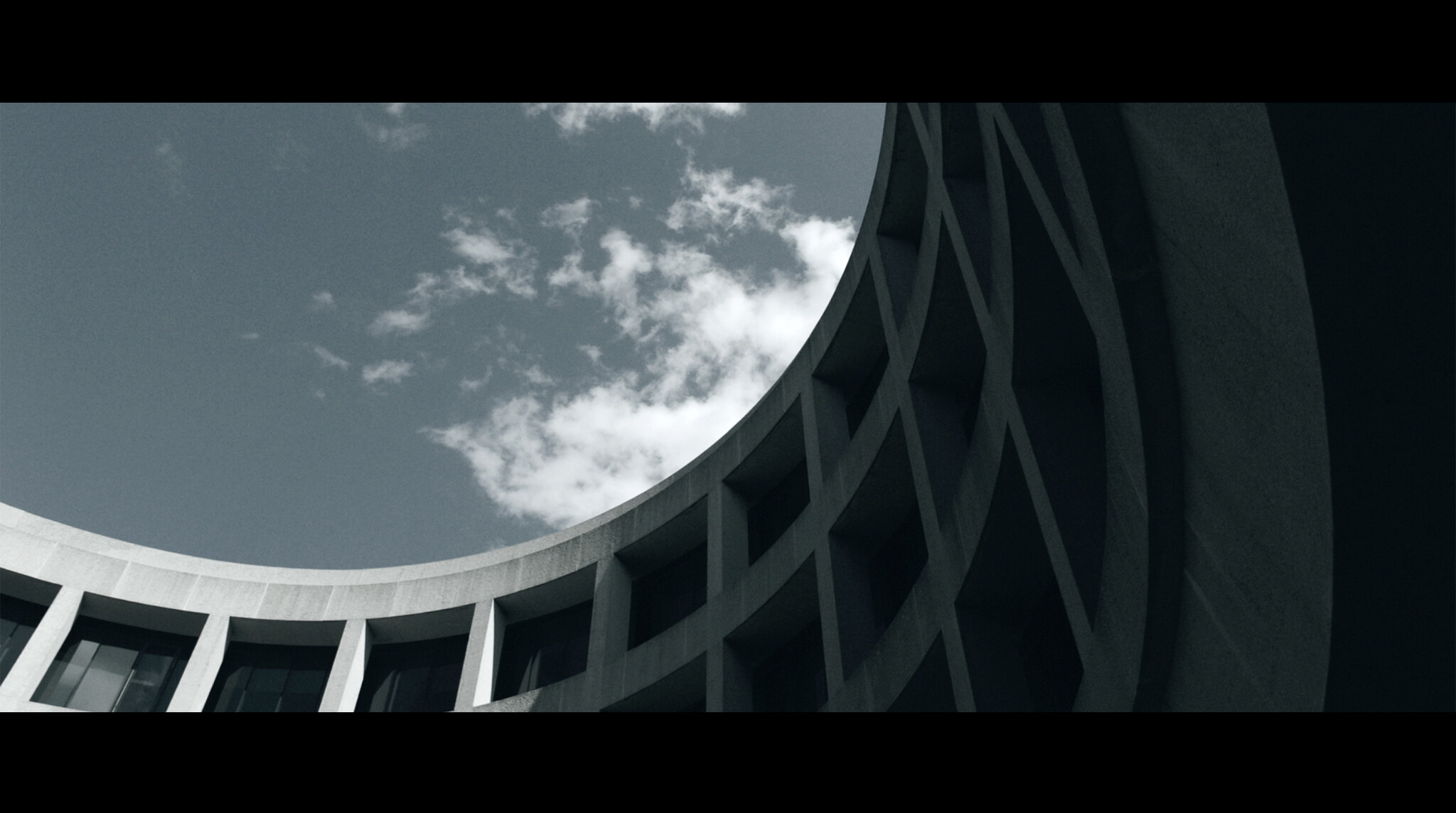 Curved concrete building façade with repeating window openings framed against a partly cloudy sky.
