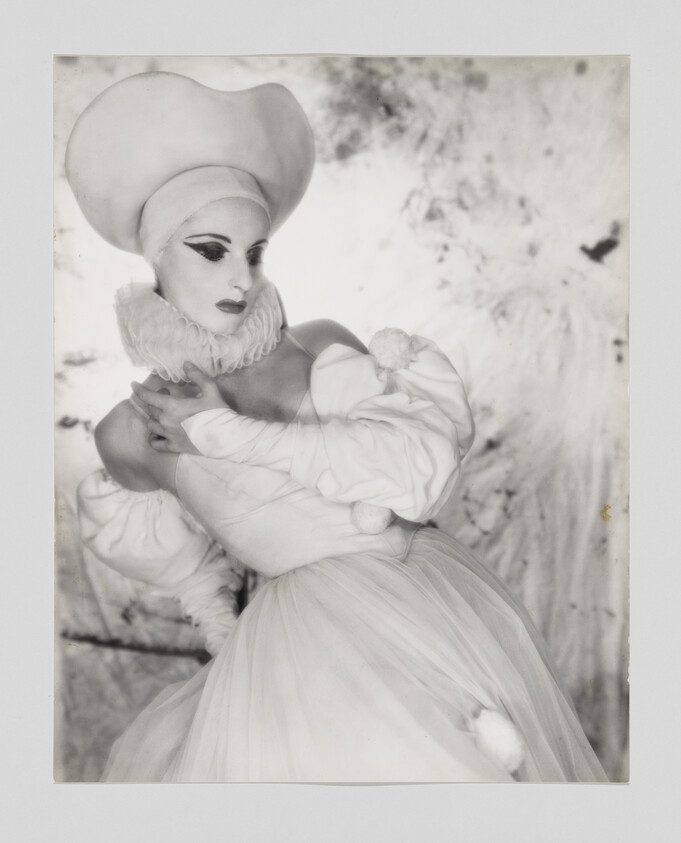 A black and white photograph of a person dressed in an elaborate costume with a large, puffy hat and ruffled collar, reminiscent of a historical or theatrical outfit. The individual is posing with their arms crossed, and their makeup and expression give off a dramatic and stylized appearance. The background is blurred and indistinct, emphasizing the subject in the foreground.