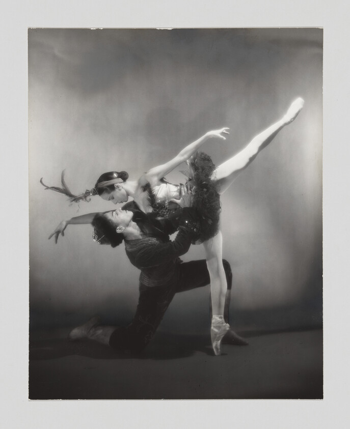 A black and white photo of two ballet dancers in a dramatic pose. The male dancer is kneeling on one leg with his back arched and head tilted back, while the female dancer, en pointe, leans over him with one leg extended high in the air. Both are wearing elaborate costumes, and the female dancer has a feathered headpiece. The background is a textured gradient, adding to the theatrical effect.