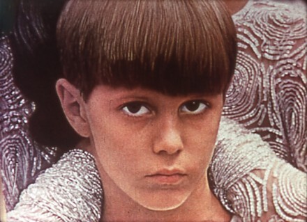 Young person with a bowl haircut looks directly at the camera with a serious expression.