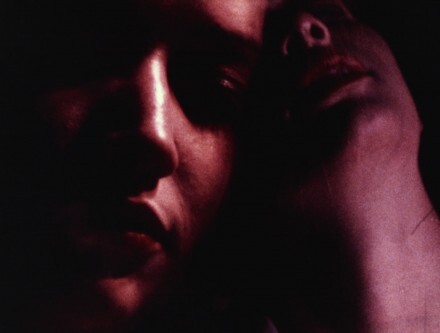 Two faces close together in dim red lighting, one leaning against the other.