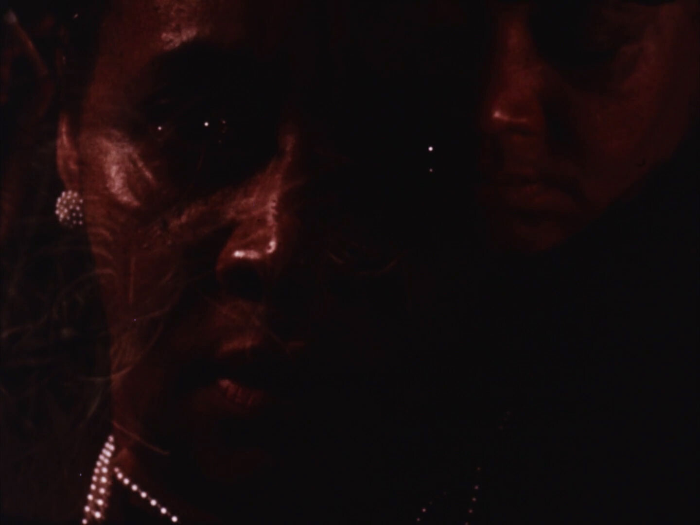 Two overlapping faces in dim red light, one wearing pearl earrings and a necklace.