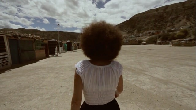 Woman with curly hair walks across a dusty open plaza toward hills and small buildings.