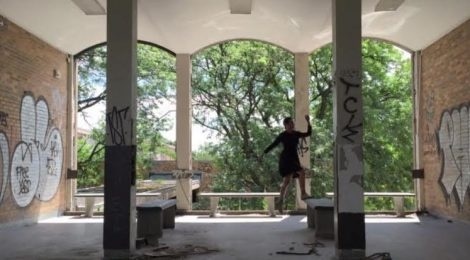 Person standing in a graffiti-covered abandoned building with open archways and greenery outside.