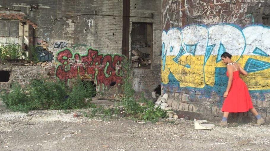 A woman in a bright red dress walks past a large wall with colorful graffiti in an abandoned area.