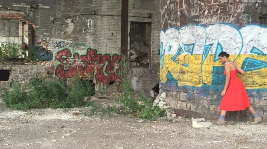 Woman in red dress walking by a graffiti-covered wall in a dilapidated urban setting.