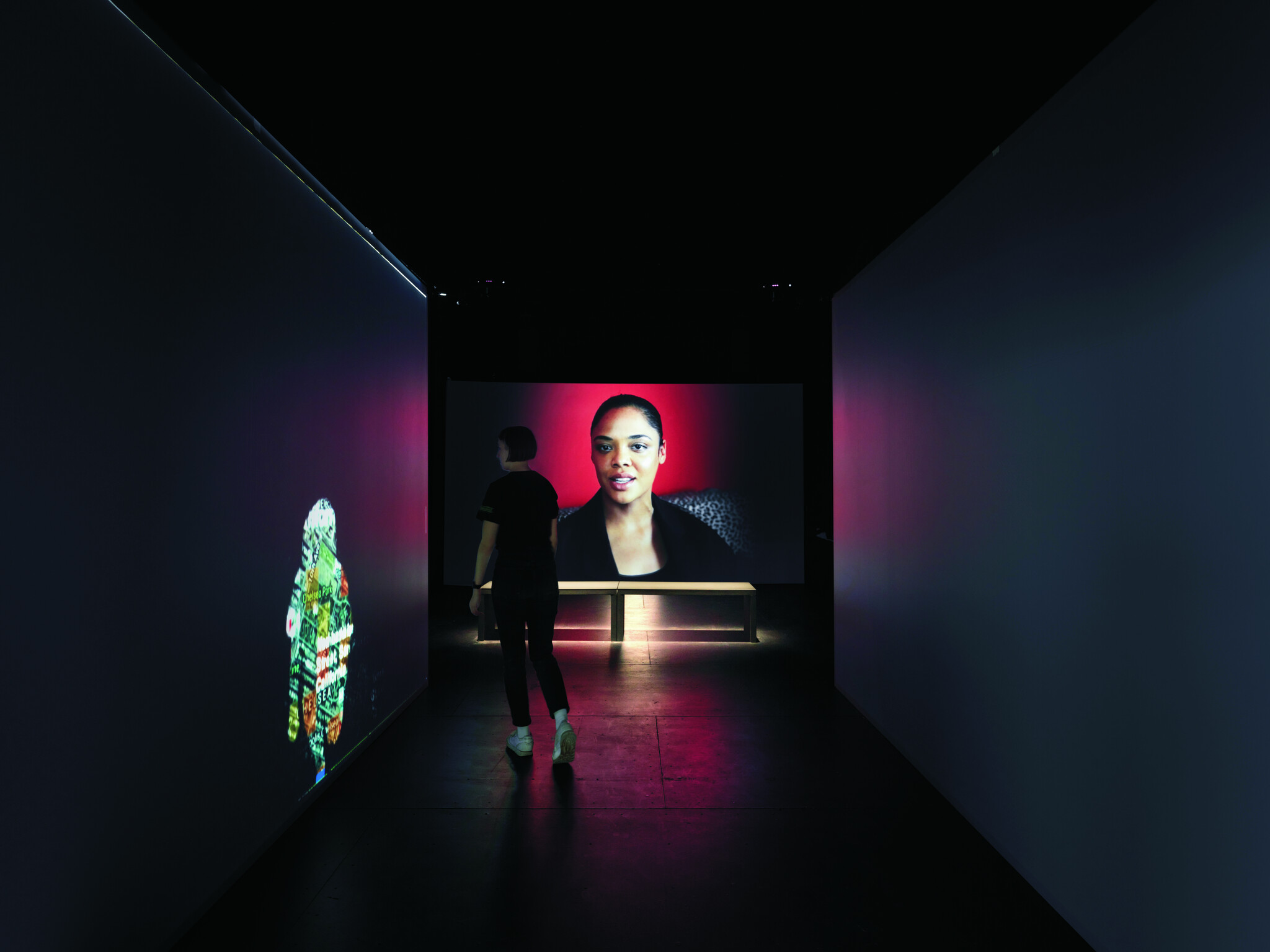 A person stands in a dark gallery space, observing a large video screen displaying a close-up of a woman's face. To the left, a colorful projection of what appears to be a fish is cast on the wall, adding a vibrant contrast to the otherwise dimly lit room.