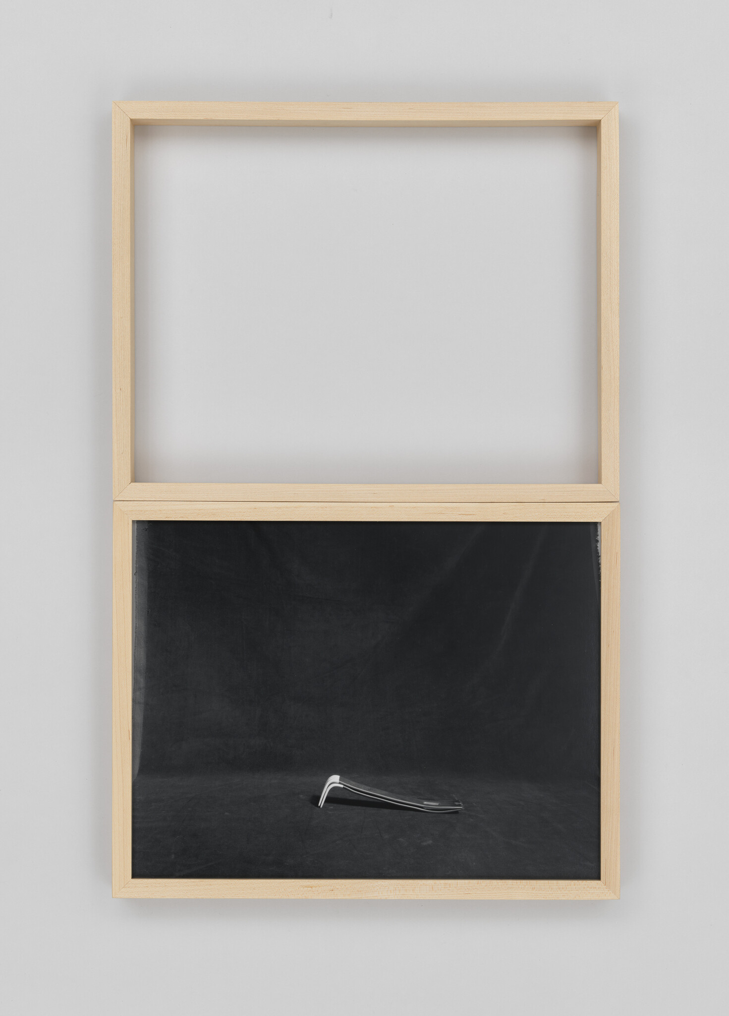 A minimalist artwork featuring a wooden frame with the top half empty and the bottom half containing a blackboard with a single silver crowbar lying diagonally across it. The background wall is white, providing a stark contrast to the blackboard.